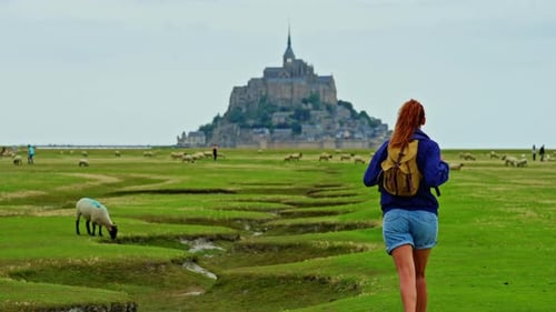 A Female with a Backpack Visits the Mont Saint Michel Castle While Walking Through Green Meadows