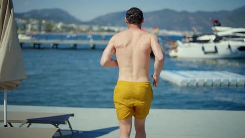 Mann in gelben Shorts springt von einem Dock mit Booten im Hintergrund ins blaue Meer