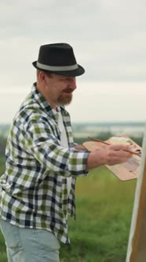Focused Painter in Hat and Checkered Shirt Working on Canvas in a Grass Field