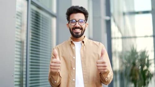 Young smiling bearded man showing thumb up looking at camera while standing on the street near