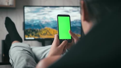 a young guy looks at the green screen of the phone and swipes his finger. man resting at home with a