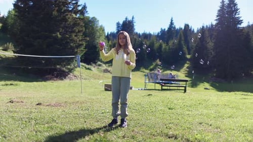Girl in Bright Suit Plays Outdoors and Blows Soap Bubbles on Sunny Meadow in Backyard
