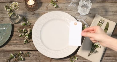 Stylish table setting. Woman putting card, floral decor and burning candle on background, above view