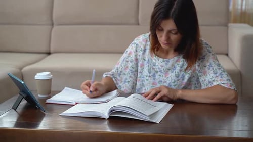 A woman watches a video on a tablet and makes notes in her notebook