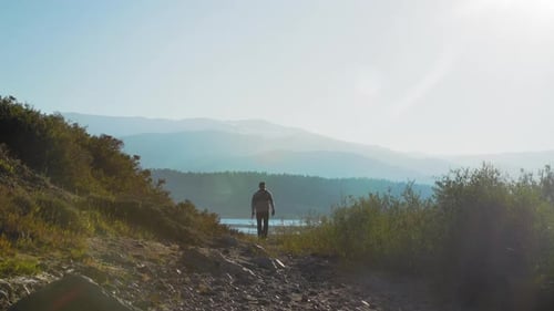 Plan large d'un pêcheur s'éloignant d'un lac en randonnée. Le soleil illumine sa forme, projetant une silhouette
