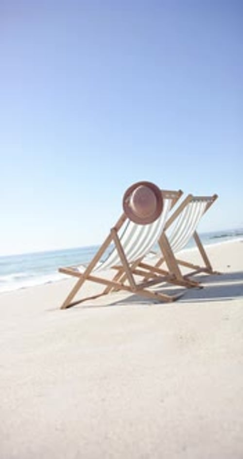Vertical video: Beach scene with two empty lounge chairs and hat, relaxing by sea