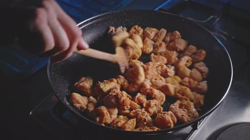 Mixing floured and seasoned pieces of chicken in a frying pan using a wooden spoon, close up view.