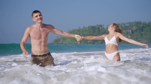 Young Fit Couple Holding Hands Having Fun Among the Waves in the Ocean Water Splashes and Foam