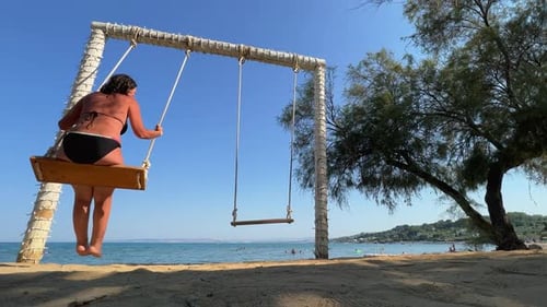 Low angle back view of woman on summer vacation enjoying relaxing and swinging on rope swing while l