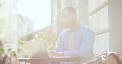 Handsome Young Man Sitting at Table of Cafe in City Center with Laptop and Typing on Keyboard