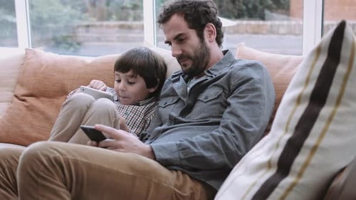 Man and child relax on couch using devices