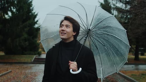 Front View of a Young Man in a Coat Walking Under an Umbrella During a Rain in the Autumn City Park