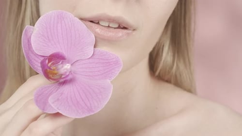 Female Lips with Delicate Pink Lipstick and Orchid Flower Closeup on a Pink Blurred Background A