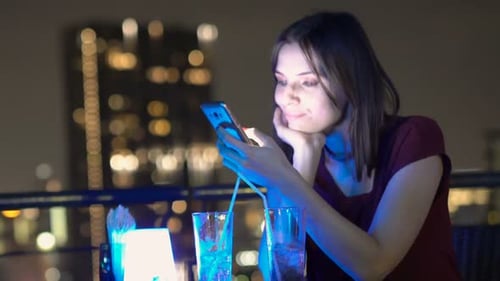 Young woman smiles browsing internet on smartphone at night in city rooftop bar