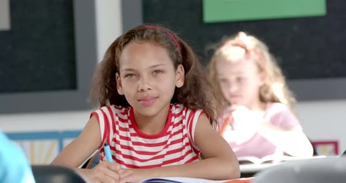 Caucasian girl smiles at the camera, biracial girl focuses on her work in a classroom in school