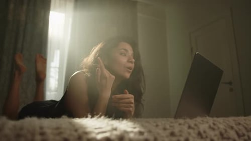 Süße Frau, die per Videoanruf auf Laptop kommuniziert und auf dem Bauch auf dem Bett liegt