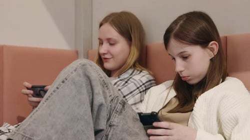 Zwei Mädchen im Teenageralter, die auf einem Bett liegen und in ihrem Gerät versunken sind, eines mit einem Smartphone, das andere mit einem