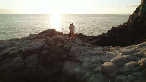 A couple embraces on rocky terrain at Los Gigantes Cliffs in Tenerife, with the setting sun casting