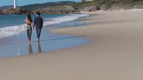 View Behind Tourist Couple Strolling Barefoot At Serene Beach With Lighthouse In Distance. Muxia, Sp