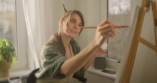 Chica morena concentrada sonriendo y pintando en caballete mientras practica pintura en casa