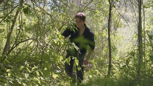 Tourist Kneels to Open Bag While Changing Direction in Dense Forest