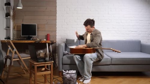 Young Man on a Sofa Practicing Acoustic Guitar While Following an Online Lesson on His Laptop in a