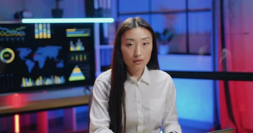 Asian woman which working on computer in evening office and then looking into camera with friendly