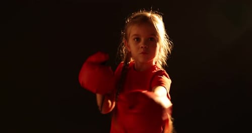 Cute little girl in protective gloves having boxing practice on black background, camera moving in