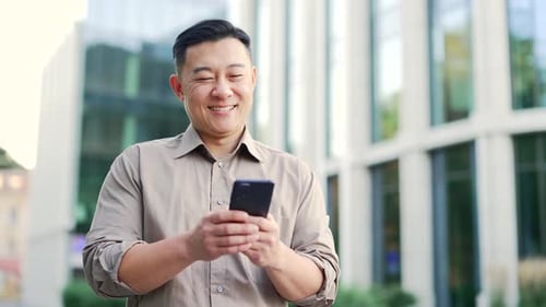 Smiling businessman in shirt is using a smartphone while standing on street near an office building.