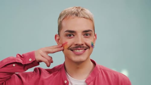 Front Shot of Happy Young Blonde Man Drawing Gay Rainbow Flag on Cheeks in Studio