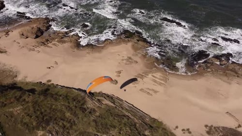 Aerial tracking shot of Paraglider with orange parachute flying over beautiful coastline and beach i