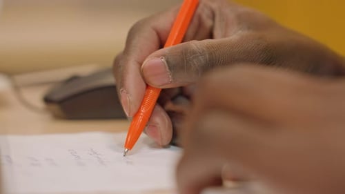 Closeup Hands Filling Exam Answers on Paper Orange Pen Tip Mouse in Background Focused Student