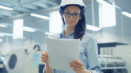 Female Engineer Wearing Hard Hat Works With Blueprints while Standing in the Middle of Big Factory.