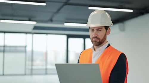 Confident Engineer in Orange Vest and White Helmet Smiles While Working on Laptop in Empty Office