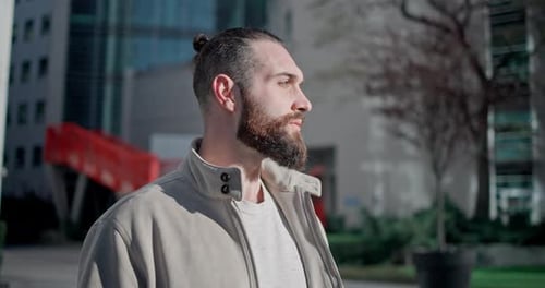 A Young Man with Stylish Trendy Hairstyle and Beard in Casual Clothes Stands on the Street in the