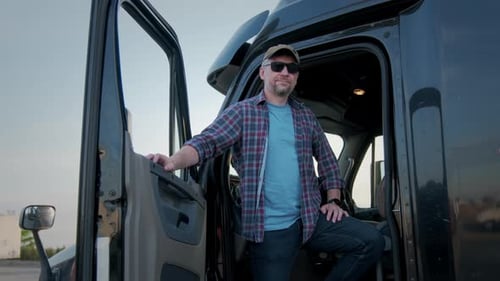 Portrait of Professional Truck Driver Sits in His Truck and Behind Him Parked Long Haul SemiTruck