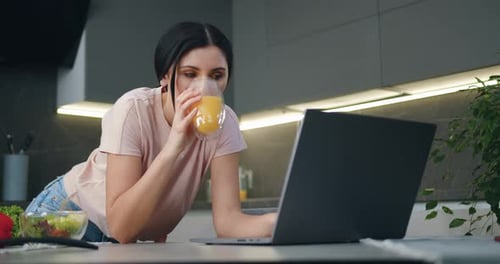 Woman Uses Laptop, Drinks Juice in Kitchen