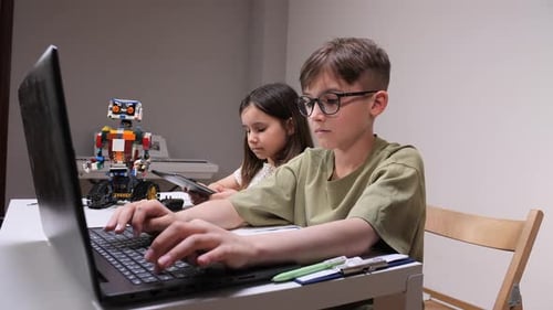 Young Boy and Girl Concentrate on Tablet and Laptop While a Colorful Robot Stands Behind Them Hands
