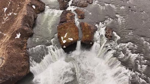 Luftaufnahme von Wasser, das über felsiges Gelände mit Schneeflocken in einer Naturlandschaft rauscht