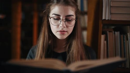 Frontansicht einer Studentin mit Brille, die das Buch in der Bibliothek liest und die Seite umblättert