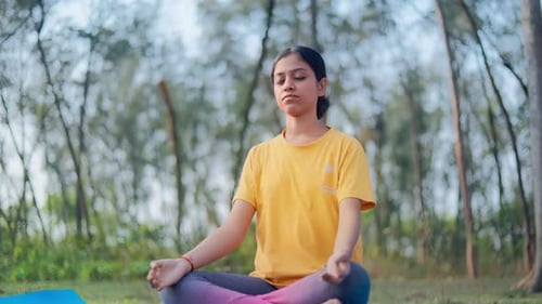 Peaceful young woman sitting in lotus position on yoga mat in the forest.