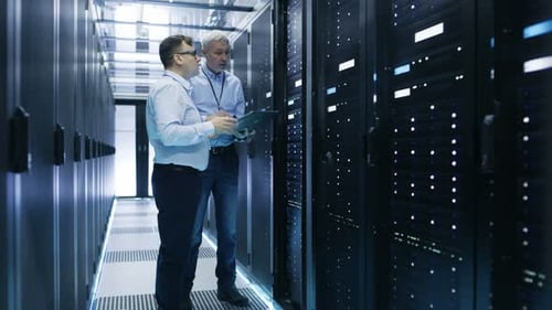 Two IT Engineers Standing in Working Data Center. They Use Laptop and Tablet Computer while Standin