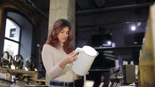 A Cute Girl with Beautiful Hair Examines a White Electric Kettle in a Home Appliance Store