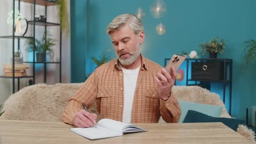 Mature Man on Phone Writing at Wooden Desk