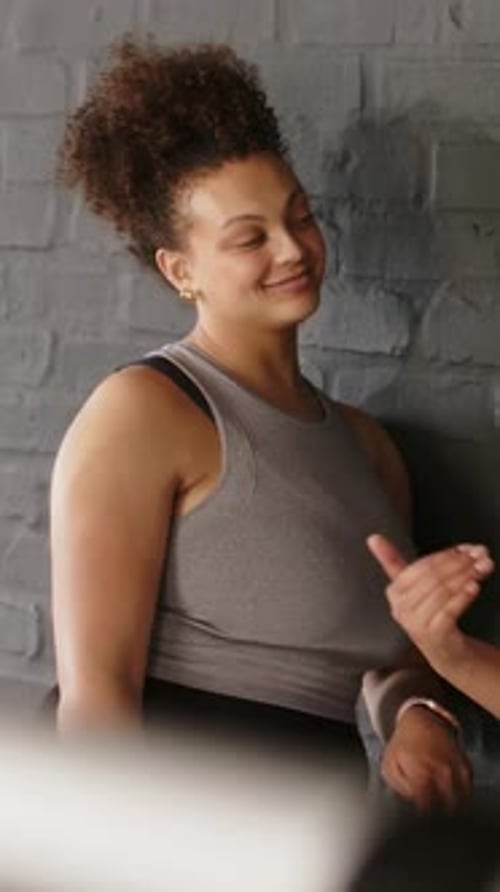 Vertical video: Responding to offframe hand gestures, woman in gray tank listening at brick wall