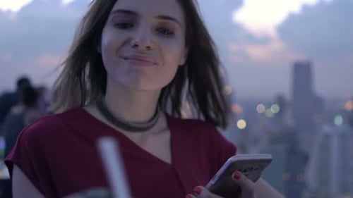 Young woman sipping cocktail uses smartphone on luxurious city rooftop terrace