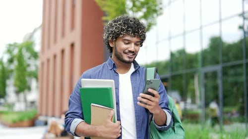 A male student uses a mobile phone while walking in campus space near university building. Handsome