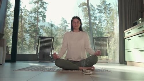 Woman Meditating in Lotus Position in Modern Home with Forest View
