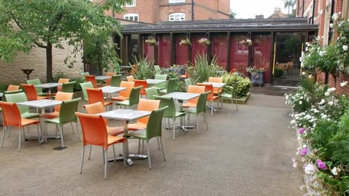 An outdoor café area featuring a mix of orange and green chairs around square tables. The space is