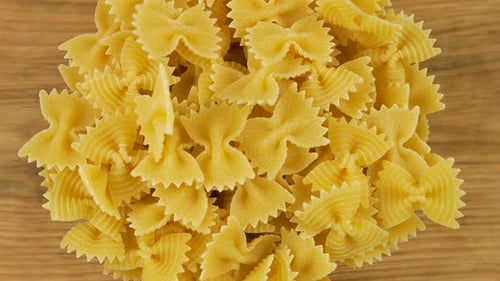 Bow-shaped pasta is poured into a glass transparent bowl against an oak wooden board. Top view. Slow